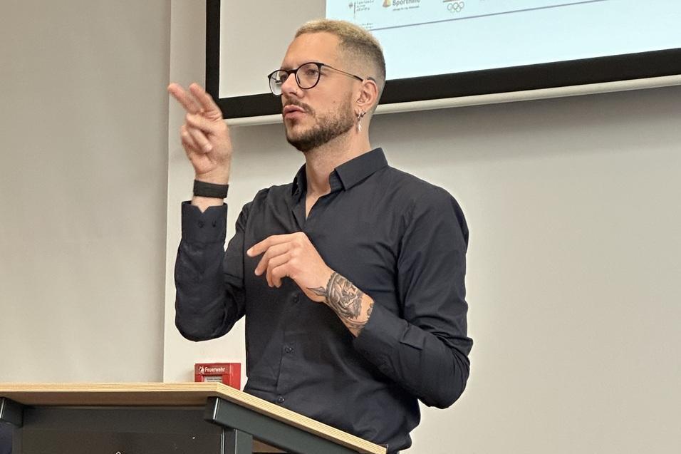 Mann mit Brille und Tätowierung spricht gebärdensprachlich an einem Rednerpult in einem Konferenzraum.