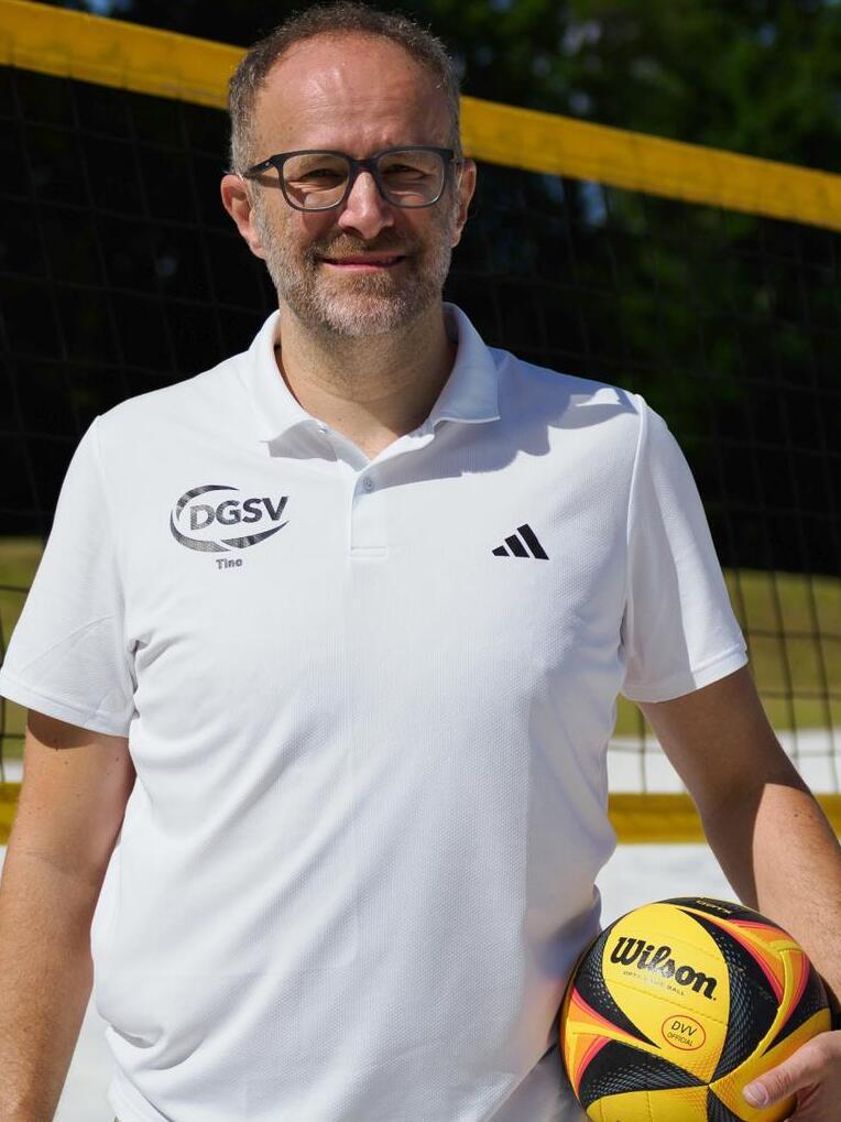 Mann in weißem Poloshirt hält einen Volleyball vor Netzhintergrund im Freien.