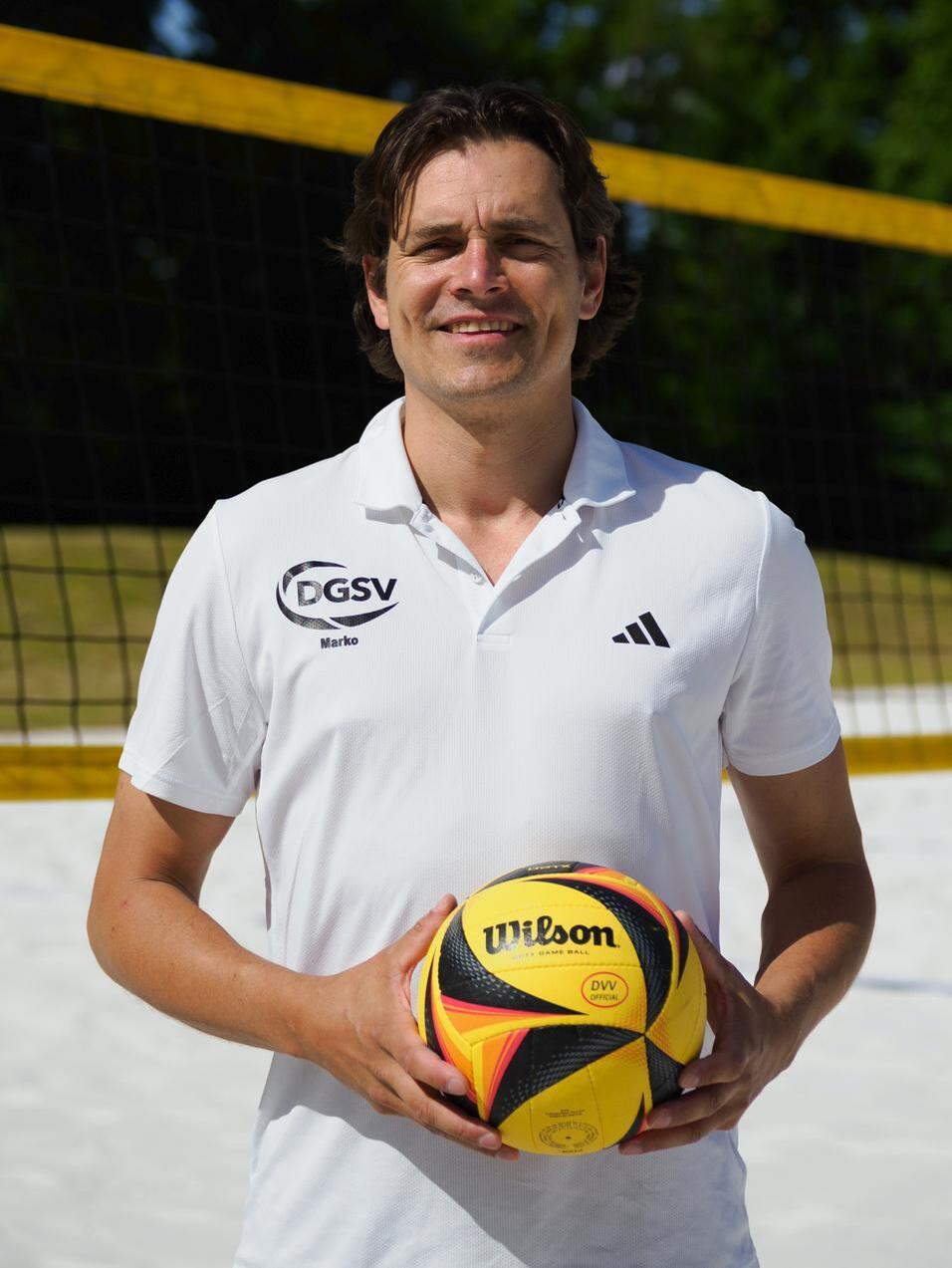 Mann in weißem Sportshirt hält einen bunten Volleyball vor einem Beachvolleyballnetz im Freien.