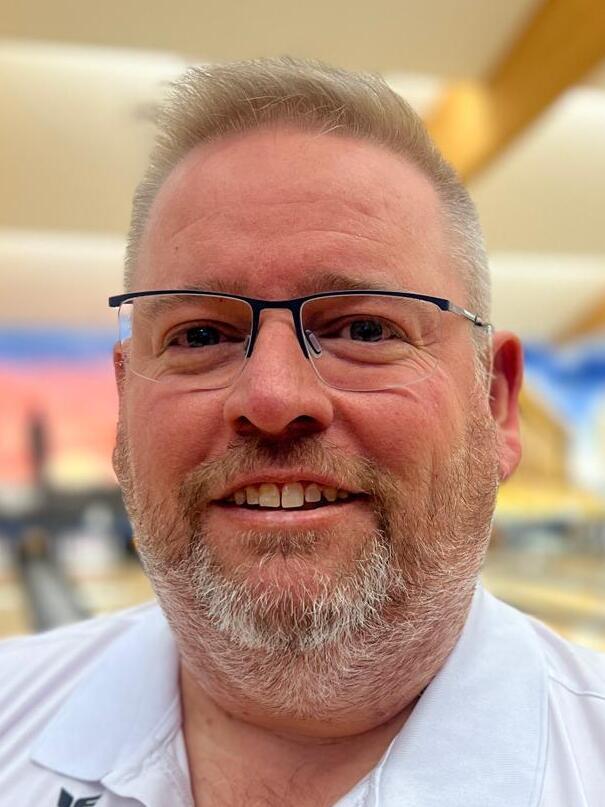 Lächelnder Mann mit Brille und Bart, vor einer farbigen Bowlingbahn.