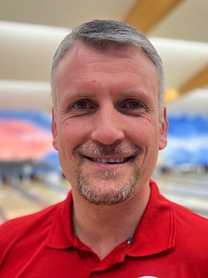 Person mit kurzem grauem Haar und Bart, lächelnd in rotem Shirt, vor einer Bowlingbahn.