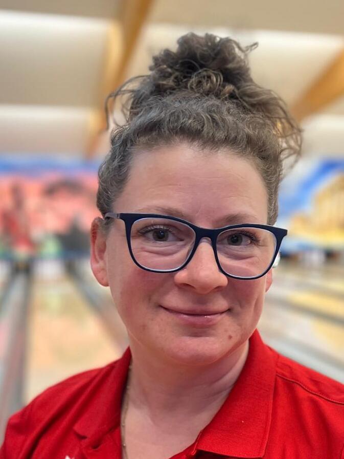 Porträt einer lächelnden Frau mit Brille, in rote Kleidung, mit Bowlingbahn im Hintergrund.