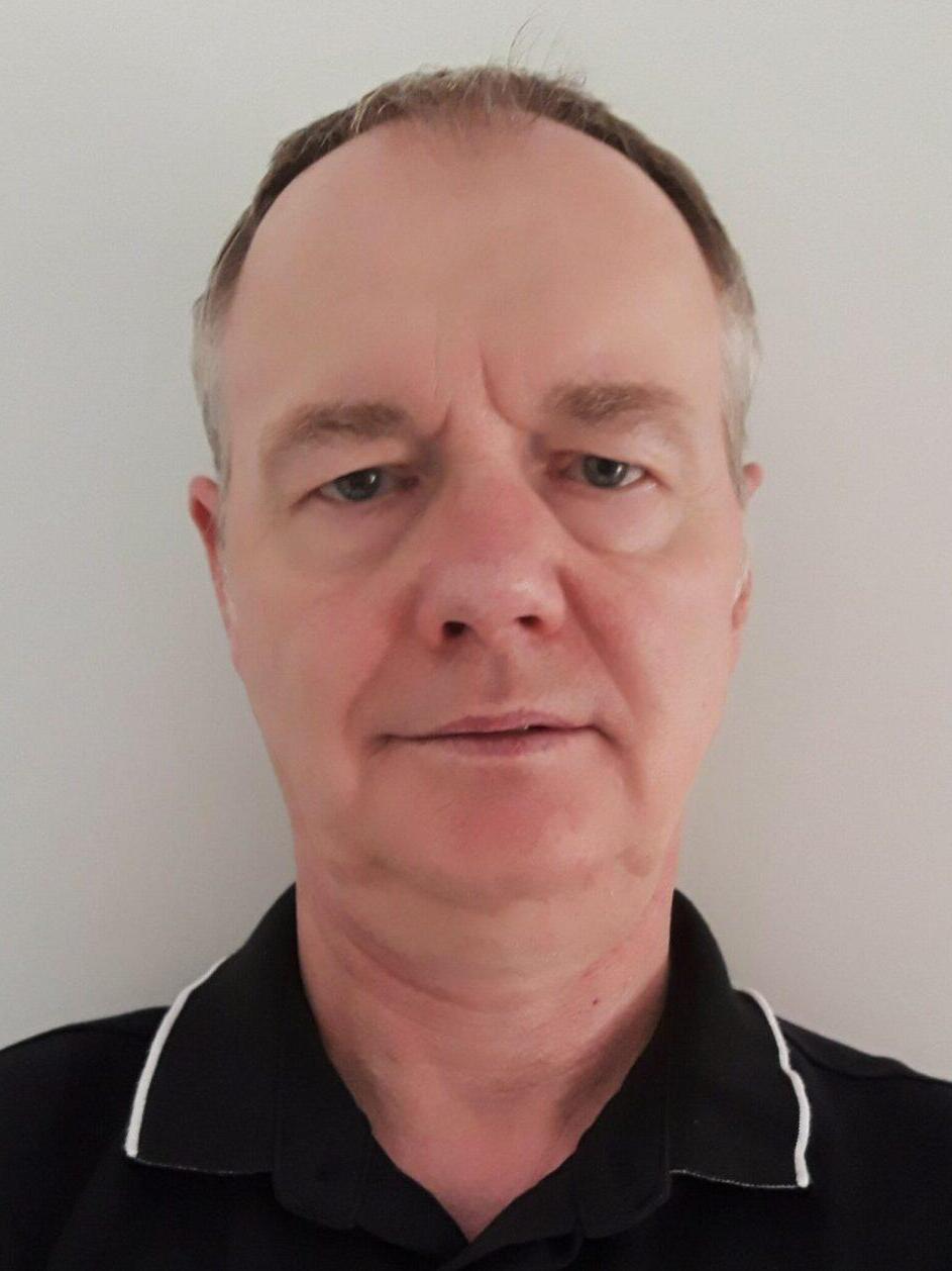 Porträt eines Mannes mit kurzen, grauen Haaren, der ein schwarzes Poloshirt trägt. Hintergrund ist hell und einfarbig.