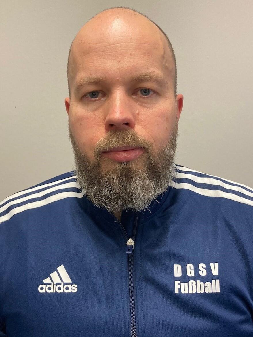 Porträt eines bärtigen Mannes in blauer Trainingsjacke mit Logo des DGSV Fußball vor grauem Hintergrund.