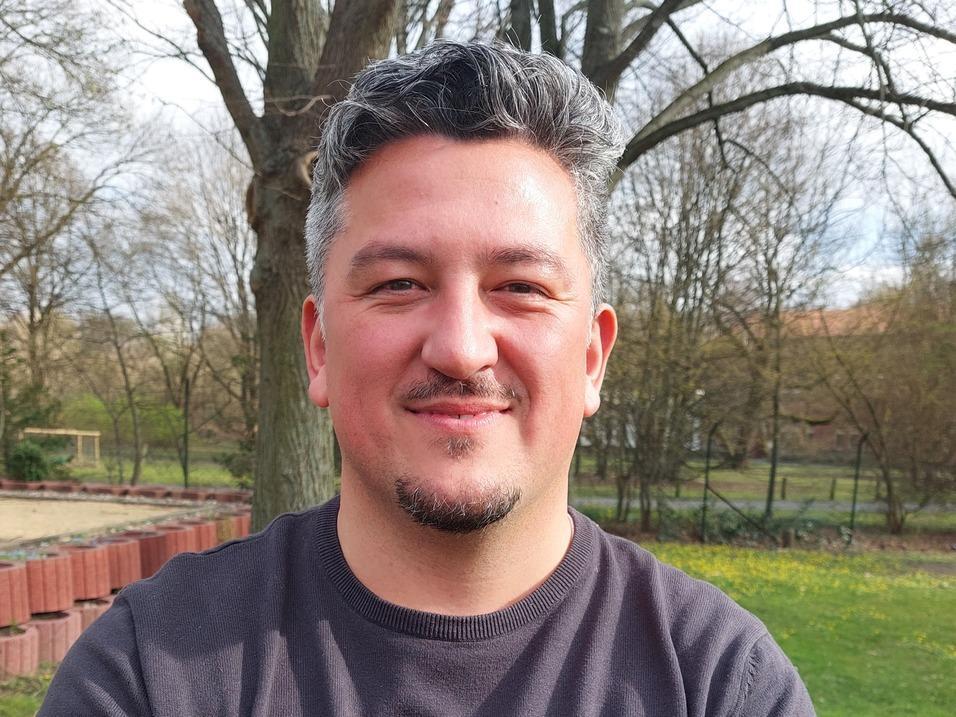 Lächelnder Mann mit grauen Haaren steht im Freien vor Bäumen und blühendem Gras.