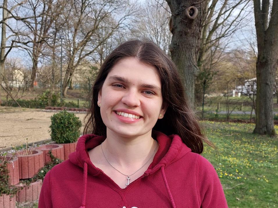 Junge Frau mit langen Haaren lächelt vor einem Baum in einem Park. Grüne Wiese und Blumen im Hintergrund sichtbar.