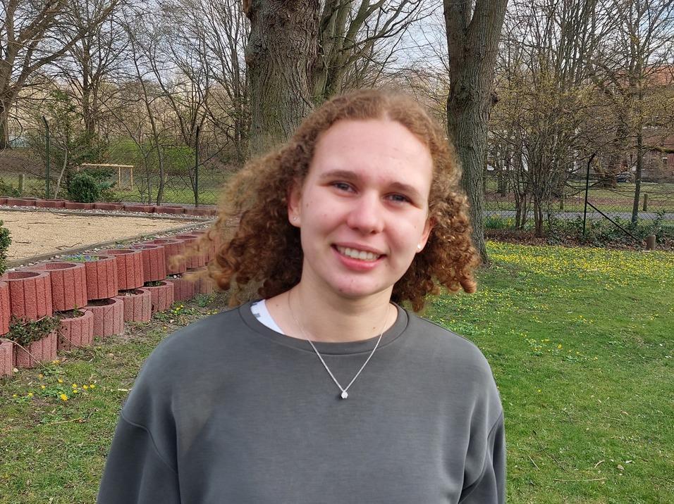 Junge Frau mit lockigem Haar lächelt vor einem grünen Gelände mit Blumen und einer roten Mauer im Hintergrund.