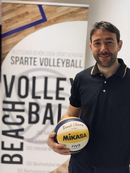 Mann hält einen Beachvolleyball der Marke Mikasa vor einem Banner der Sparte Volleyball.