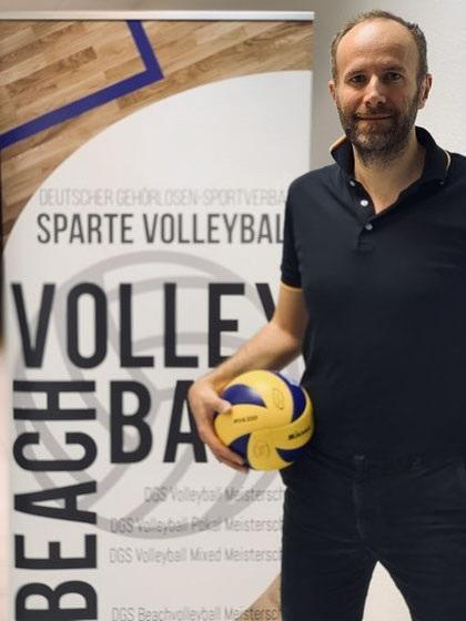 Mann hält einen Volleyball in der Hand, posiert vor einem Banner der Sparte Volleyball des Gehörlosen-Sportverbands.