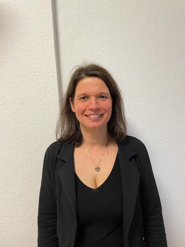 Silke Porträt einer Frau mit braunen Haaren, die in einem schwarzen Outfit vor einer weißen Wand steht.