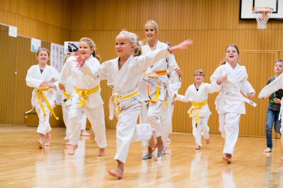 Gruppe von Kindern in weißen Karateanzügen mit gelben Gürteln, die fröhlich in einer Sporthalle herumrennen.