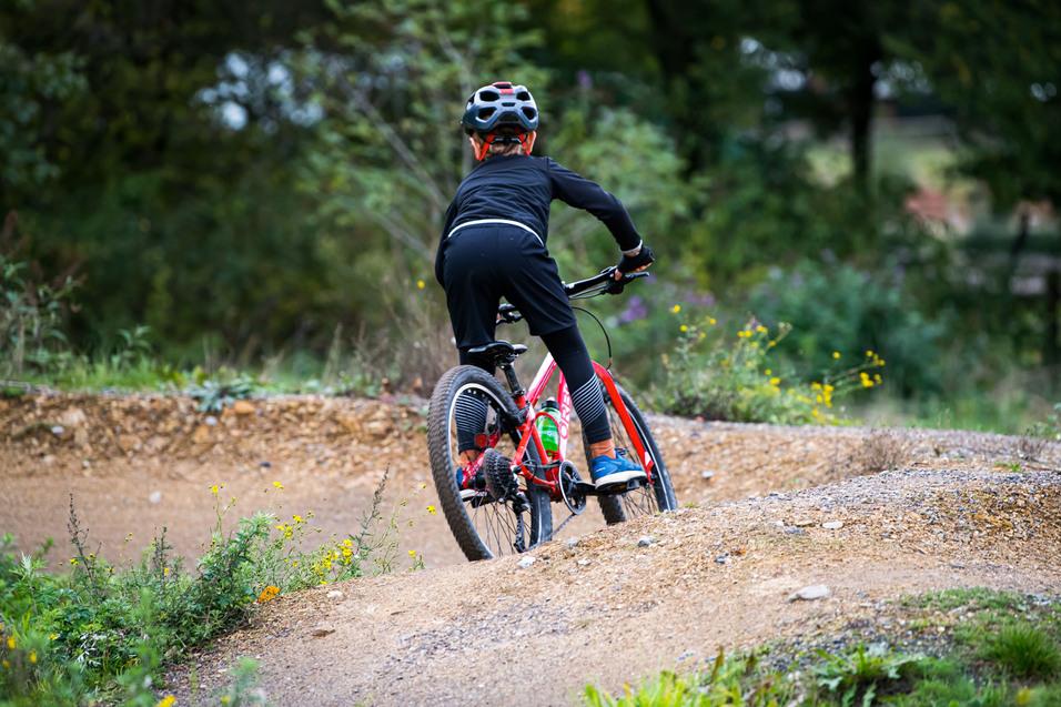 Ein Kind fährt mit einem roten Mountainbike über eine kurvige, gravelige Strecke in der Natur.
