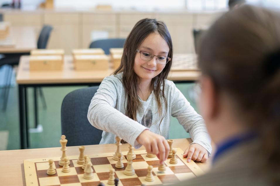 Mädchen mit Brille zieht eine Schachfigur auf einem Schachbrett in einem Klassenraum. Holzschachfiguren im Vordergrund.
