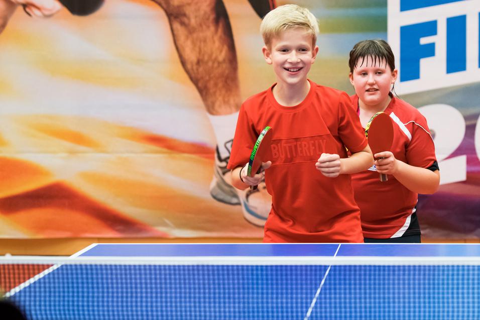 Zwei Kinder in roten Trikots stehen lächelnd an einem Tischtennistisch mit Schlägern in den Händen.